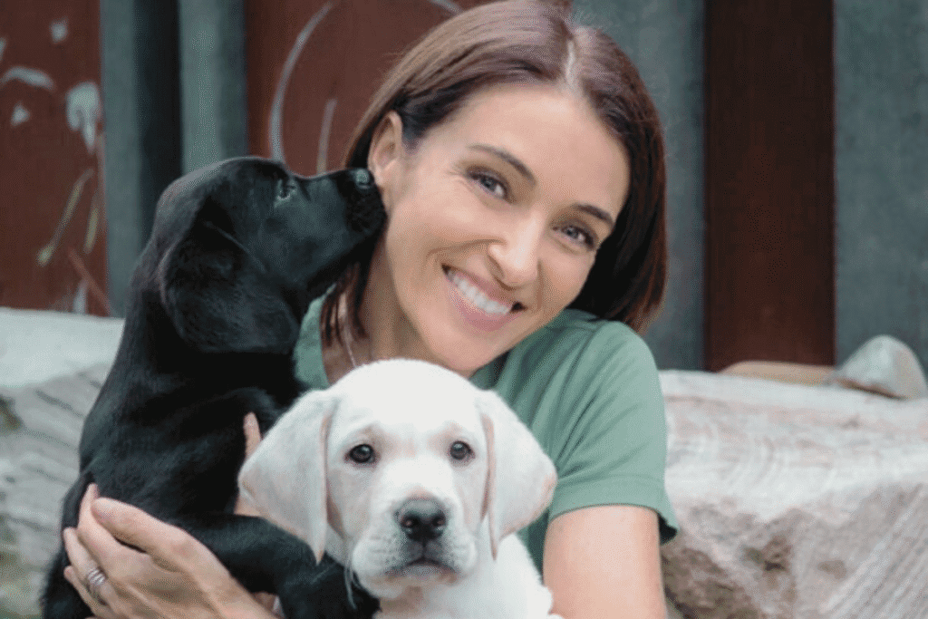 Television vet Dr Katrina Warren smiling at camera holding Guide Dog NSW puppies a white and black Labrador 